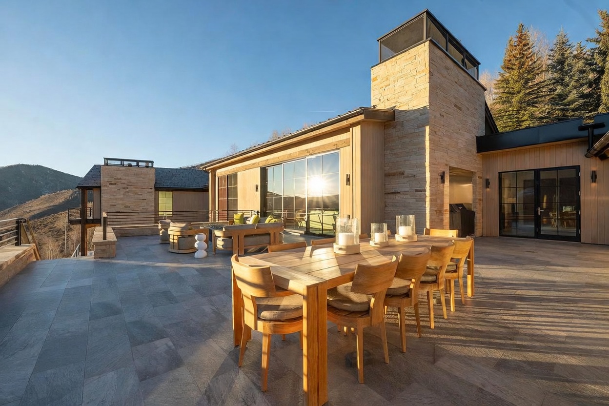 This expansive stone-paved terrace offers a luxurious outdoor living space with a dining area featuring a light wood table and matching chairs. A separate lounge area with comfortable seating is positioned near a tall stone-clad chimney breast, all set against a backdrop of mountain views and evergreen trees. The scene is captured from a slightly elevated perspective, highlighting the seamless transition between the indoor living space and the outdoor deck through large glass sliding doors.
