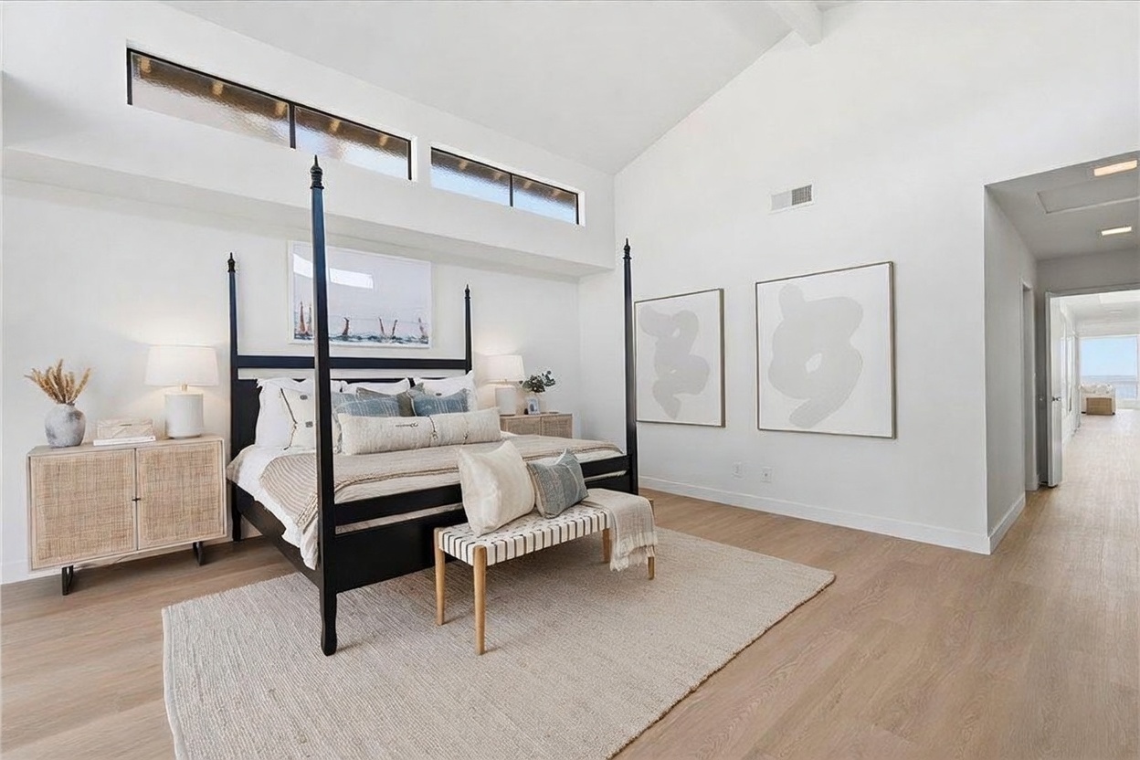 This spacious primary bedroom features a high vaulted ceiling with clerestory windows that flood the room with natural light. A prominent black four-poster bed serves as the focal point, complemented by light wood flooring, a textured area rug, and a matching bench at the foot of the bed. The minimalist decor, neutral color palette, and clean lines create a serene and sophisticated atmosphere.
