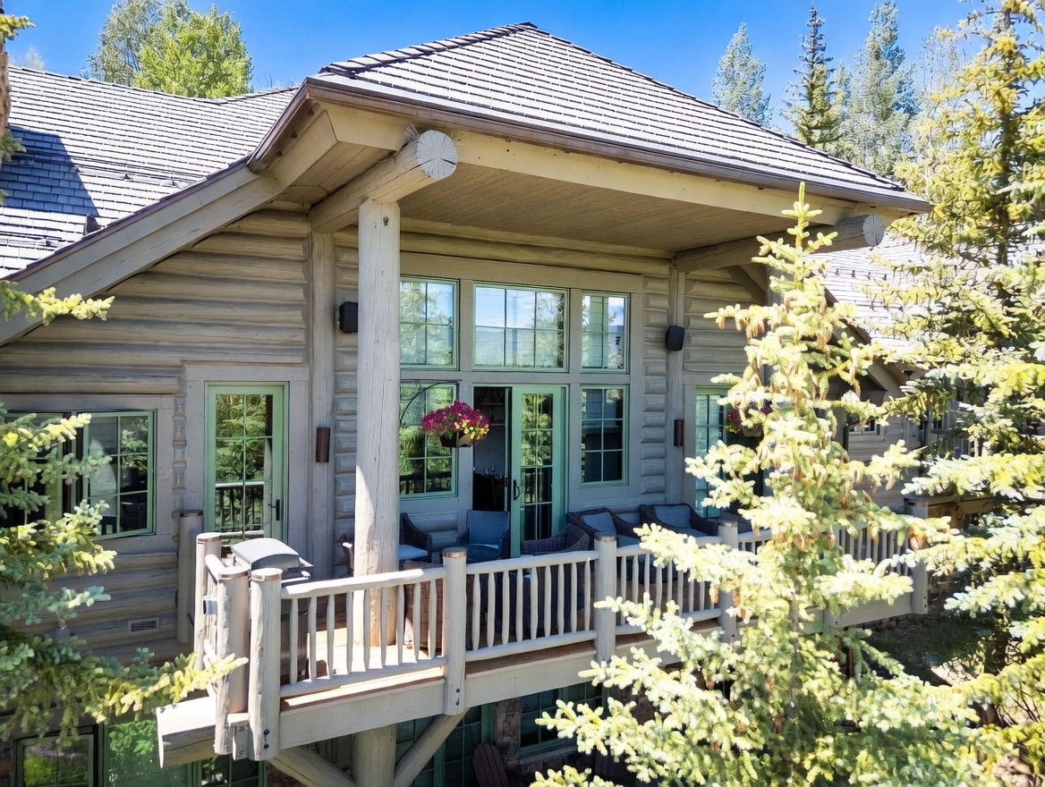 This image captures a rustic log cabin-style home from an elevated perspective, focusing on a spacious wooden deck. The deck features a railing, outdoor seating, and a grill, set against a backdrop of large windows and a covered porch area. The surrounding evergreen trees and natural setting create a serene, mountain-lodge atmosphere.