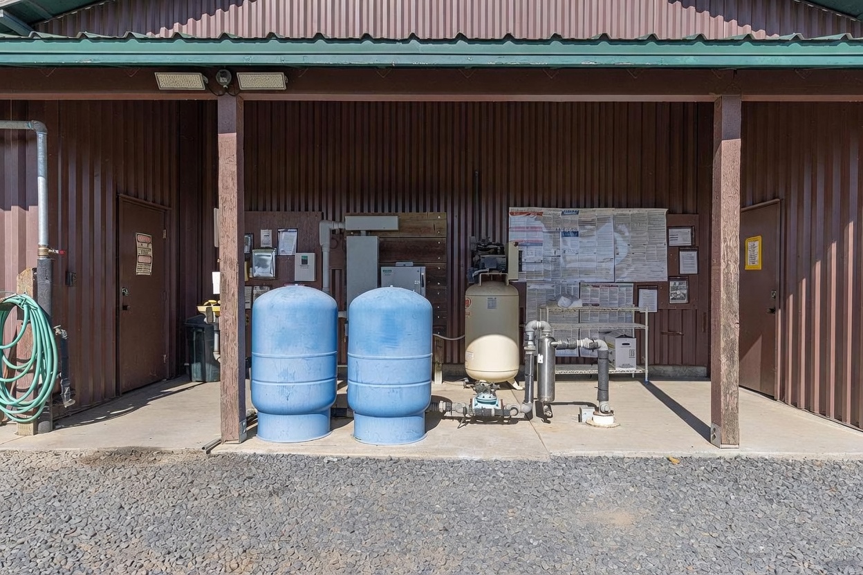 This image shows a utility or pump house structure featuring a covered, open-air bay with corrugated metal siding. The space contains industrial water filtration equipment, including two large blue pressure tanks and a beige air compressor, set against a concrete floor. A bulletin board with various documents and safety notices is mounted on the back wall, providing a functional, utilitarian aesthetic.