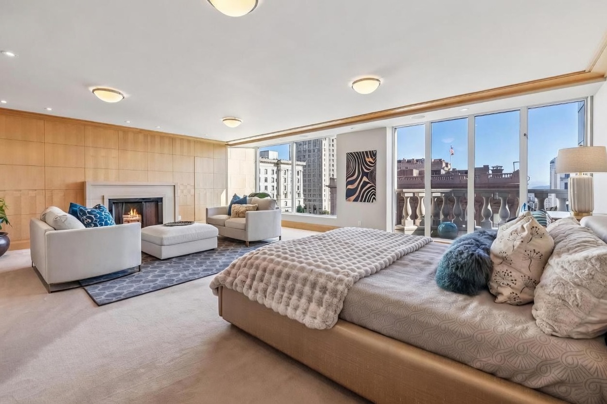 This spacious primary bedroom features a neutral color palette with light wood wall paneling and plush carpeting. The room includes a cozy seating area with a fireplace, a large bed with textured bedding, and expansive windows that provide abundant natural light and city views. The overall atmosphere is sophisticated and serene, blending comfortable living space with sleeping quarters.