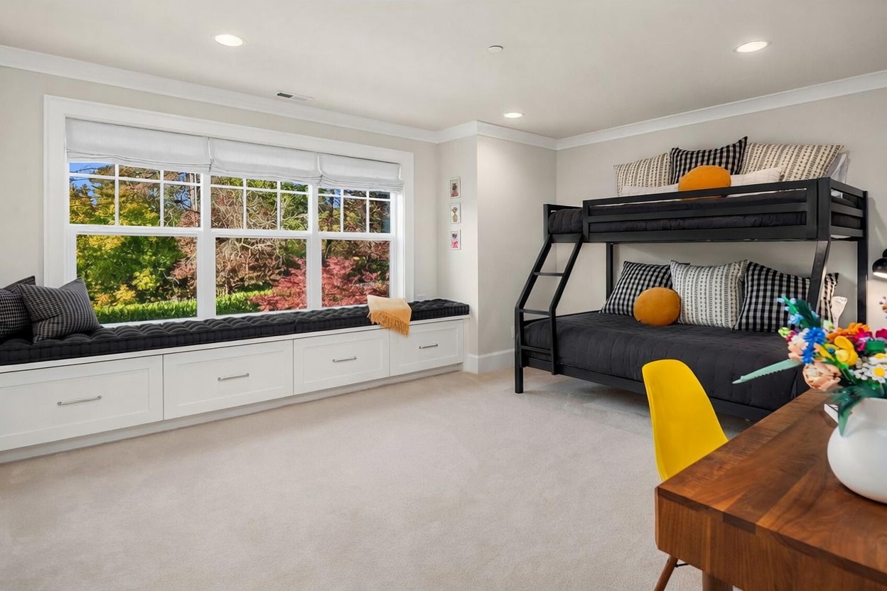 This bright and inviting bedroom features a built-in window seat with storage drawers beneath a large, multi-pane window that overlooks a lush, tree-filled exterior. A black metal bunk bed with patterned bedding serves as a focal point, complemented by a wooden desk and a vibrant yellow chair in the foreground. The room is finished with neutral carpeting and recessed lighting, creating a cozy and functional space.