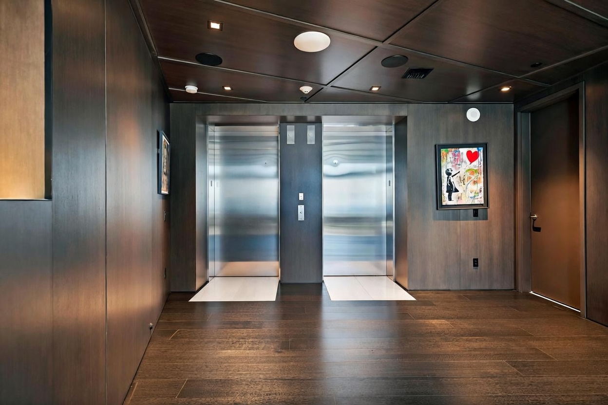 This sophisticated elevator lobby features dark wood-paneled walls and a rich, dark wood-plank floor, creating a moody and upscale atmosphere. Two brushed-metal elevator doors are centered in the space, flanked by a piece of colorful pop-art on the right wall. The ceiling is finished with a geometric grid pattern, and the overall aesthetic is modern, sleek, and highly polished.