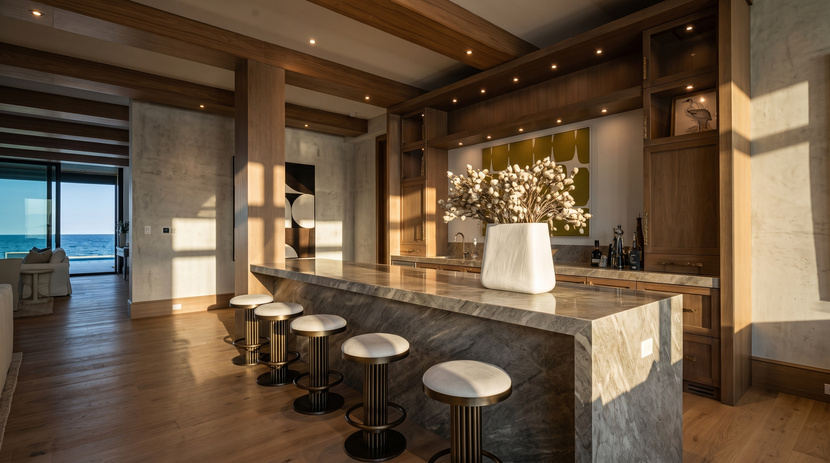This sophisticated wet bar area features a long stone-topped island with five matching metal stools, set against a backdrop of custom light wood cabinetry and integrated lighting. The space is defined by exposed wood ceiling beams and a seamless transition to an open-concept living area that leads to a view of the ocean. The overall aesthetic is refined and coastal, emphasizing high-end finishes and a bright, airy atmosphere.