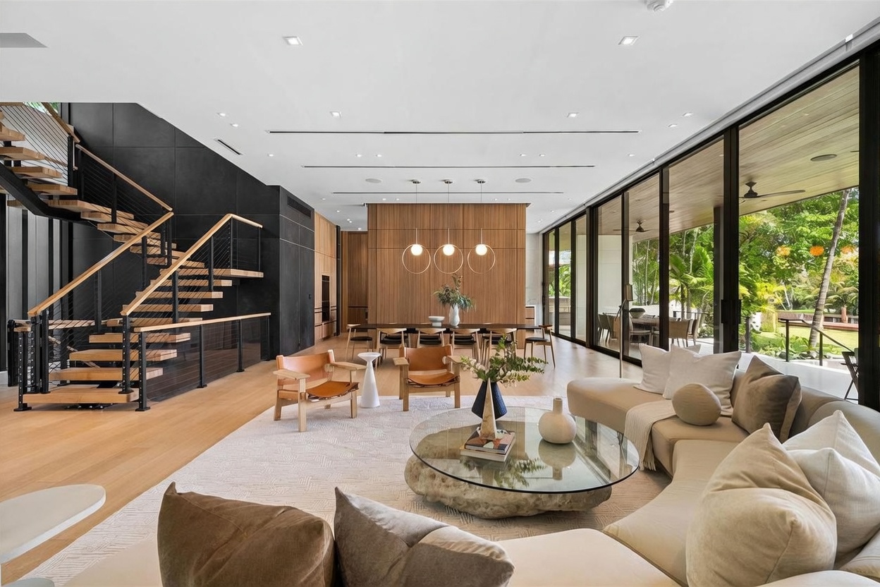 This expansive, open-concept living area features a modern aesthetic with light wood flooring and a striking black accent wall alongside a floating staircase. The space flows seamlessly into a dining area defined by a warm wood-paneled wall and three minimalist pendant lights, while floor-to-ceiling glass walls provide an unobstructed view of the lush outdoor greenery. A large, curved sectional sofa and a unique stone-based coffee table anchor the living space, creating a sophisticated and airy atmosphere.