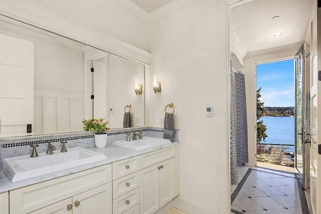 This elegant primary bathroom features a double vanity with white cabinetry, marble countertops, and a large wall-to-wall mirror. A doorway leads directly to a shower area that opens onto a balcony, offering a stunning, unobstructed view of the lake. The space is bright and airy, combining classic cabinetry with a luxurious, scenic connection to the outdoors.