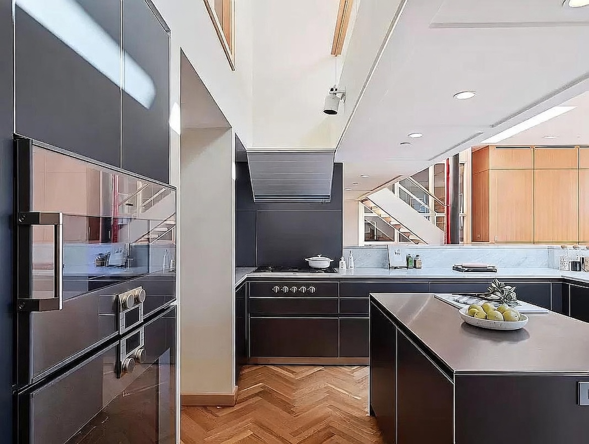 This modern kitchen features dark, sleek cabinetry paired with a large central island and a herringbone-patterned wood floor. The space includes a built-in oven, a gas range with a stainless steel hood, and a light-colored stone backsplash that contrasts with the dark finishes. The perspective captures the depth of the kitchen, looking past the island toward a staircase in the background, creating an open and sophisticated atmosphere.