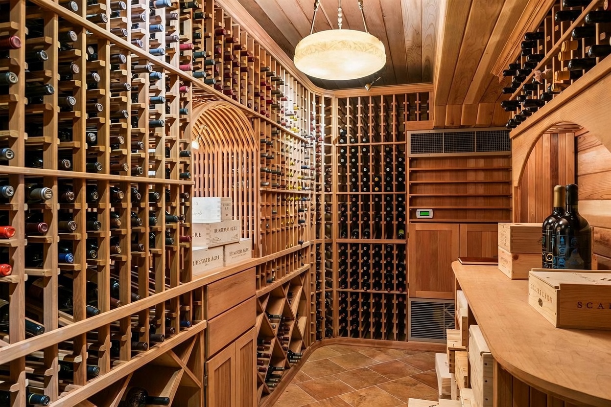 This sophisticated wine cellar features extensive custom wood racking that spans the walls, providing ample storage for a large collection. The space is illuminated by a warm, central pendant light and includes a dedicated climate control unit, with wooden crates stacked on a sturdy counter surface. The room is finished with terracotta-toned floor tiles, creating a classic and refined atmosphere for wine storage.