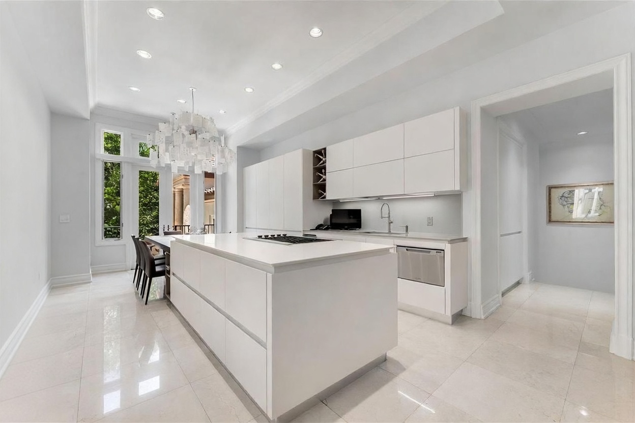 This sleek, contemporary kitchen features a minimalist design with crisp white cabinetry and a large central island topped with a smooth, light-colored surface. A striking, multi-tiered modern chandelier hangs above the island, while large windows provide a view of the outdoor greenery. The space is finished with polished light-colored flooring, creating a bright and airy atmosphere that flows into an adjacent hallway.