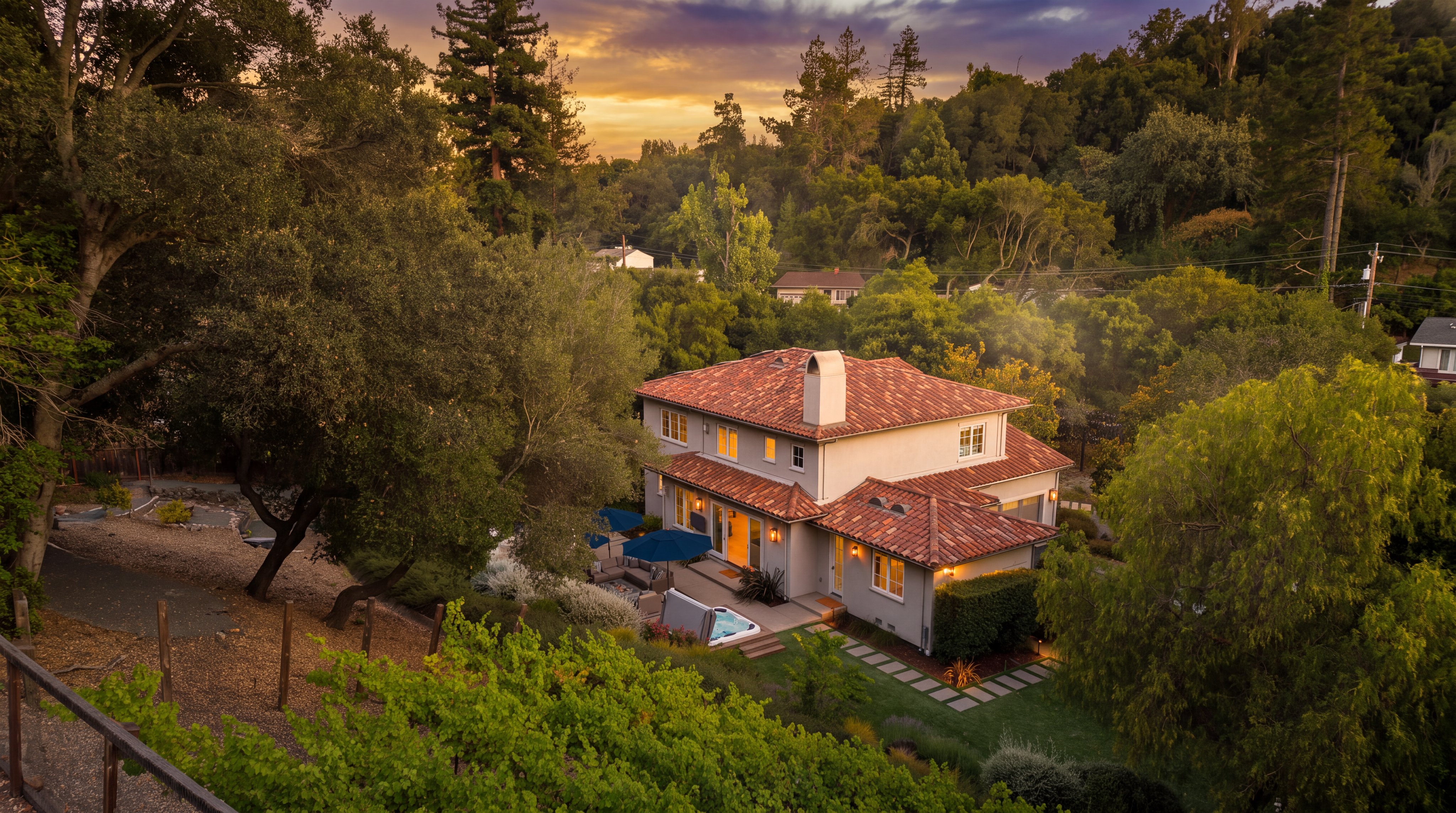 This elevated aerial perspective captures a Mediterranean-style home nestled within a lush, wooded landscape. The property features a distinctive terracotta-tiled roof, a private backyard with a patio, a hot tub, and a well-manicured lawn bordered by a vineyard. The scene conveys a serene, upscale residential atmosphere, highlighting the seamless integration of the home with its natural surroundings.