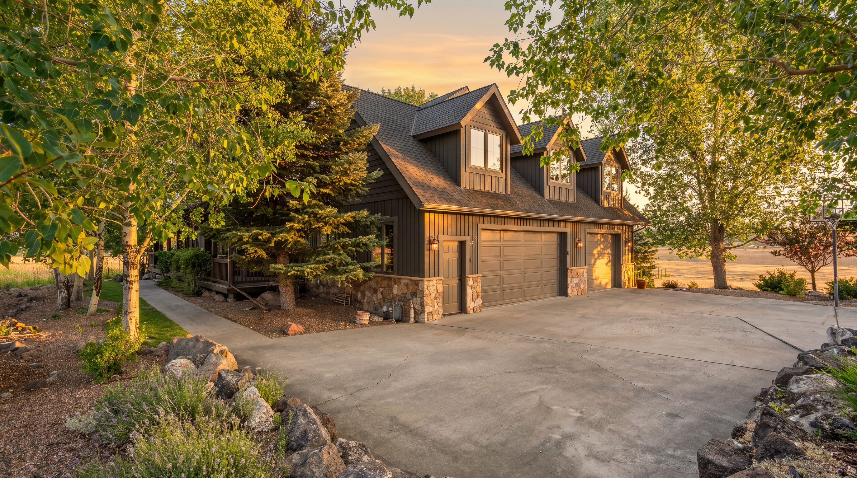 This image showcases the front-facing, multi-car garage of a custom home featuring dark vertical siding and stone foundation accents. The property includes a paved driveway, multiple dormer windows on the roofline, and mature landscaping, creating a rustic yet polished curb appeal.
