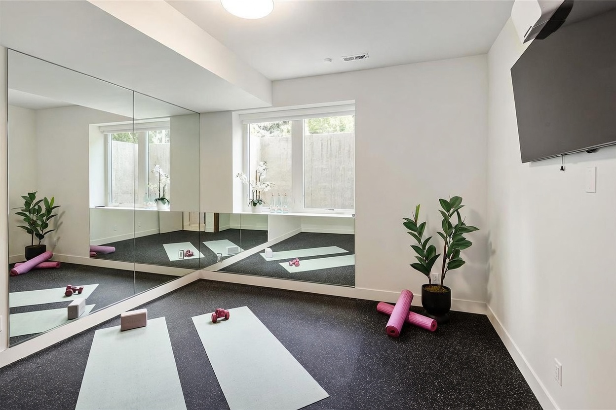 This dedicated home gym features dark, speckled rubber flooring and a large wall of mirrors that enhances the sense of space. The room is equipped with yoga mats, dumbbells, and foam rollers, while a wall-mounted television provides entertainment during workouts. A window with a view of a concrete retaining wall allows natural light to brighten the minimalist, white-walled space.