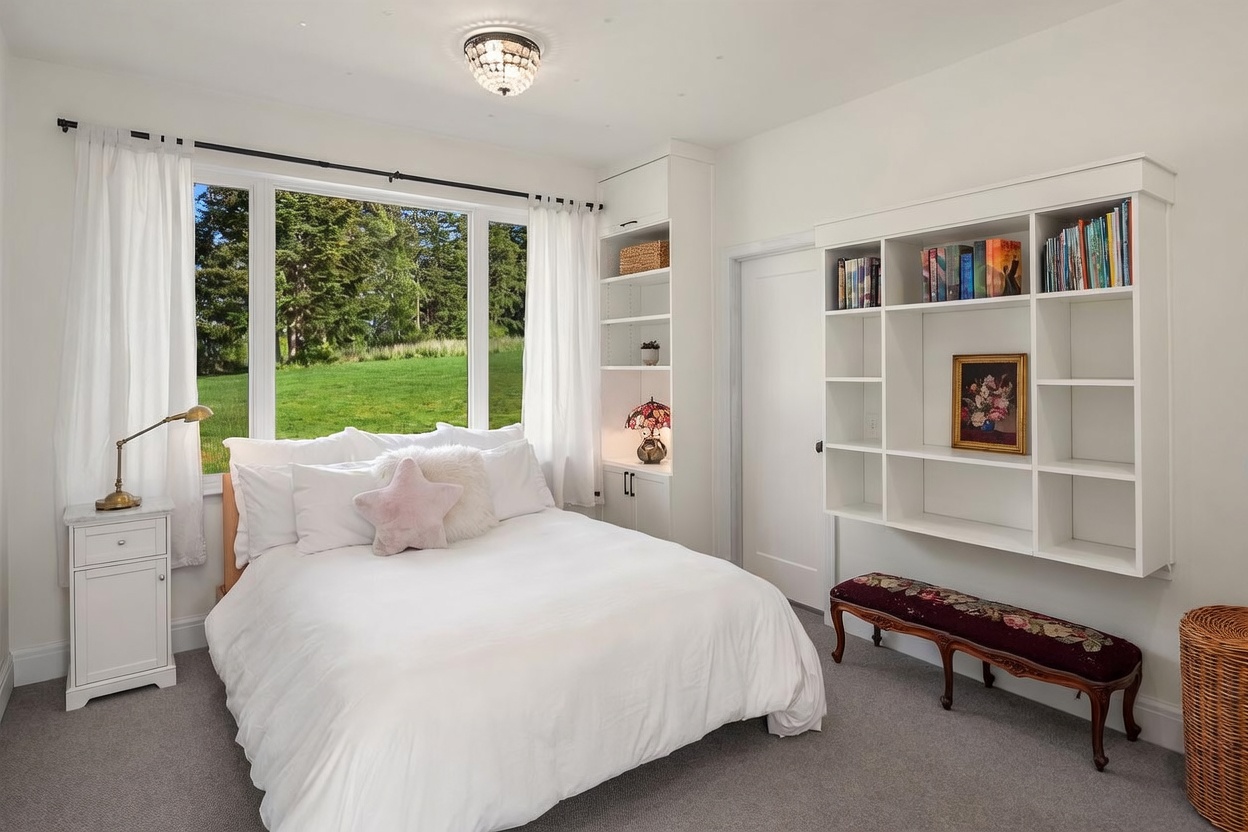 This bright and airy bedroom features a plush bed centered beneath a large window that offers a serene view of the surrounding greenery. The room is accented by built-in white shelving units, including a wall-mounted bookcase and a tall storage cabinet, providing ample display and organization space. A decorative bench sits at the foot of the bed, and a crystal-style flush-mount light fixture adds a touch of elegance to the neutral, carpeted space.