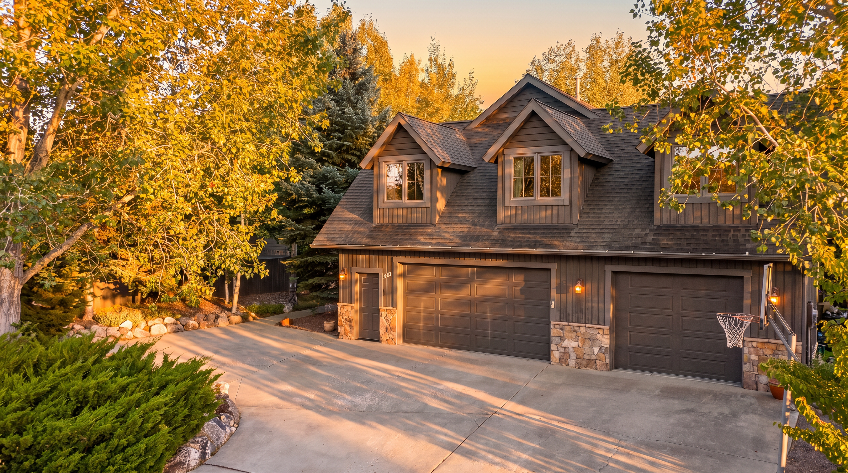 This elevated aerial view showcases a charming, rustic-style garage structure featuring dark wood siding and stone accents on a concrete driveway. The building includes two distinct garage bays, a side entry door, and two dormer windows integrated into the roof line. Surrounded by lush deciduous trees, the property offers a picturesque and well-maintained aesthetic.