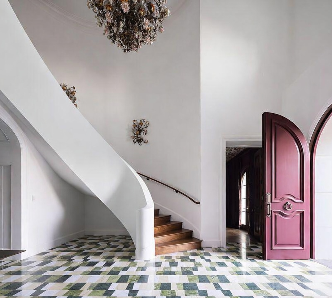 This grand foyer features a dramatic curved staircase with a smooth white balustrade and warm-toned wooden steps. The floor is adorned with a striking geometric pattern of green, white, and dark-toned tiles, while a large, ornate floral-style chandelier hangs from the high ceiling. To the right, a bold, arched magenta door stands open, revealing a glimpse into a dark-paneled room beyond.