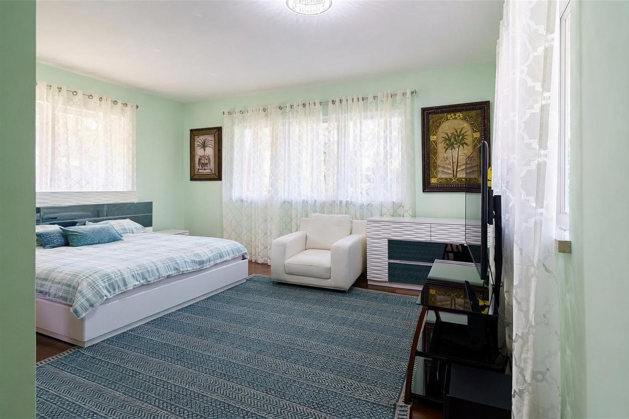 This primary bedroom features light green walls and a large, patterned area rug covering the dark wood flooring. The room is furnished with a bed dressed in blue and white plaid bedding, a white armchair, and a white dresser with textured drawer fronts. Large windows draped in sheer white curtains allow natural light to fill the space, creating a bright and airy atmosphere.