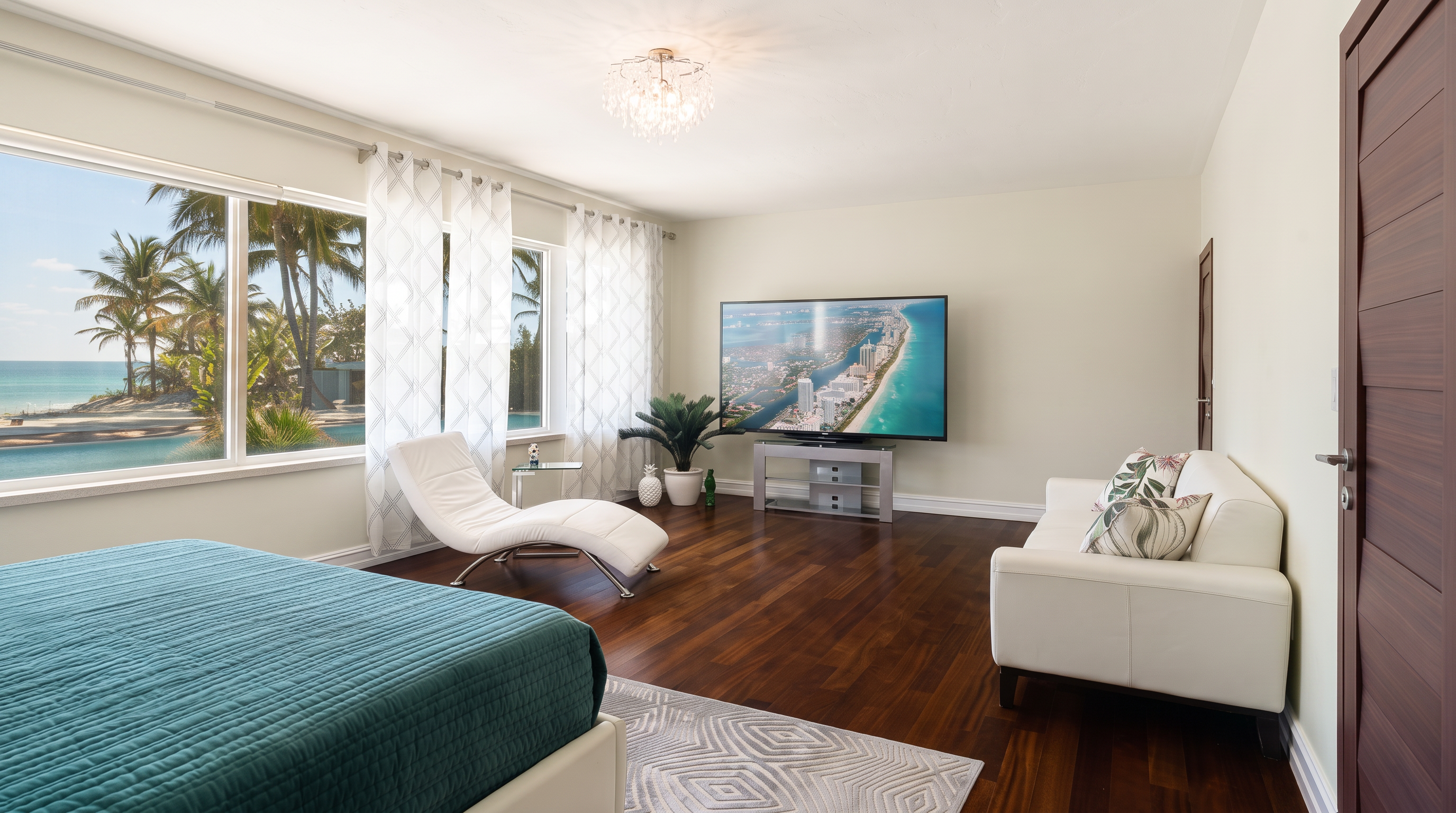 This spacious bedroom features dark wood flooring and light-colored walls, creating a bright and airy atmosphere. The room is furnished with a bed in the foreground, a white chaise lounge near the large windows, and a white sofa opposite a wall-mounted television. A crystal chandelier adds a touch of elegance, while the windows provide a scenic view of palm trees and a pool area.