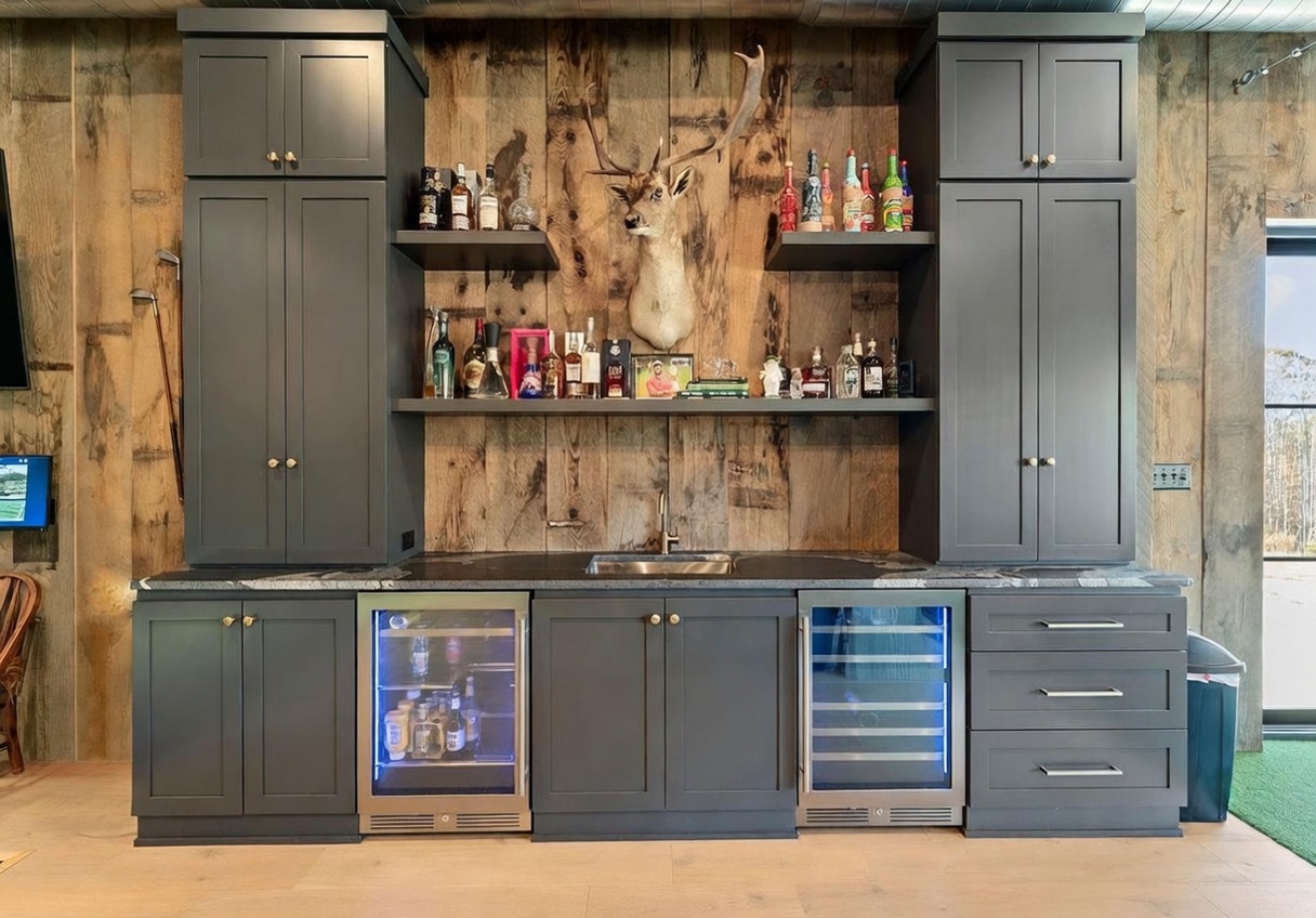 This home bar area features dark cabinetry contrasted against a rustic wood-paneled wall, creating a sophisticated, lodge-inspired aesthetic. The space is equipped with a built-in sink, two under-counter beverage refrigerators, and open shelving displaying a collection of spirits. A mounted deer head serves as a focal point above the counter, adding a distinct decorative element to the wet bar setup.