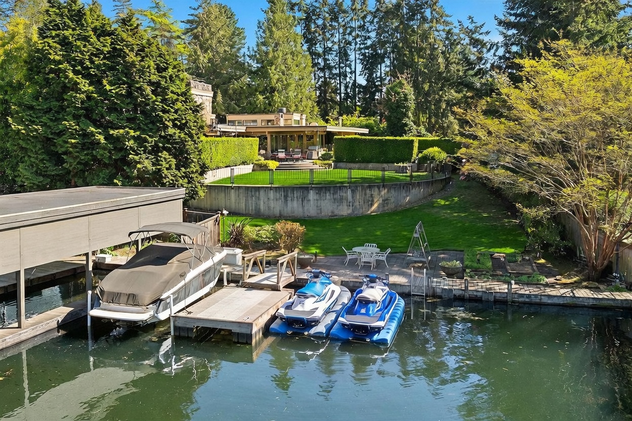 This elevated perspective captures a serene waterfront property featuring a private dock equipped with a boat and two jet skis. A lush, tiered backyard transitions from a paved patio area with outdoor seating to a manicured lawn, all enclosed by a retaining wall and mature landscaping. The main residence, visible in the background, showcases a contemporary architectural style with large windows and a flat roofline, nestled among tall, dense trees.