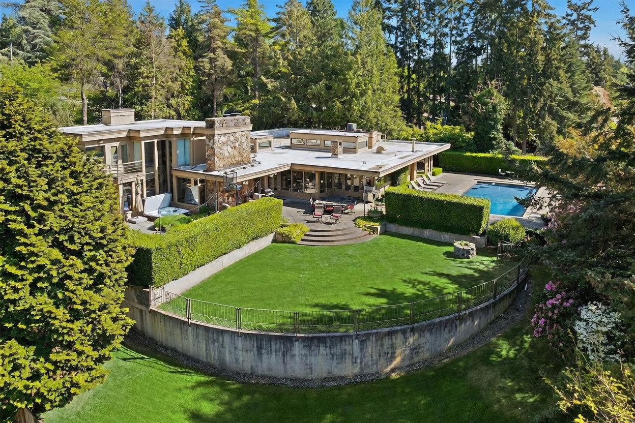 This elevated perspective captures a sprawling contemporary residence nestled within a lush, wooded landscape. The home features a flat roofline, a prominent stone-clad chimney breast, and extensive glass walls that open onto a multi-level patio area complete with a hot tub and a swimming pool. A manicured lawn is framed by a curved concrete retaining wall and mature hedges, creating a private and serene outdoor living environment.