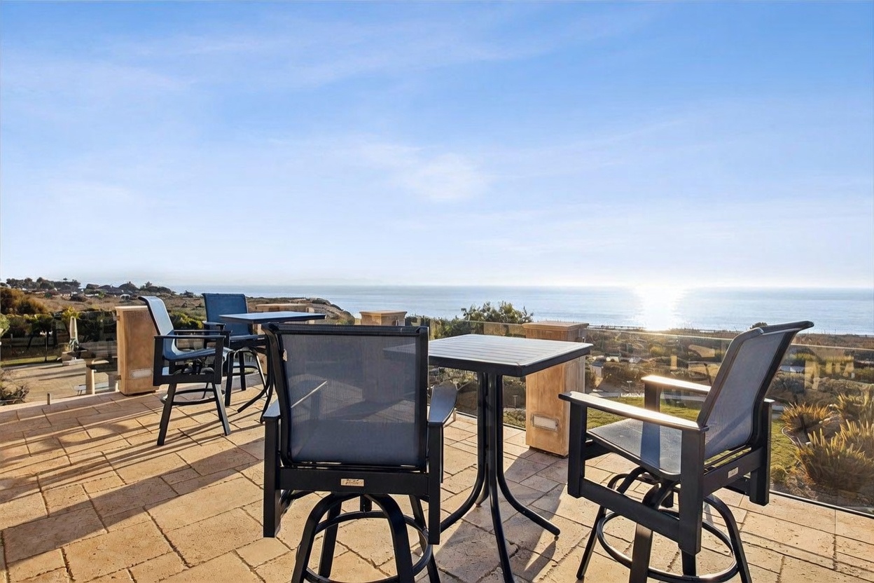 This elevated outdoor patio features stone-tiled flooring and is furnished with several dining sets, offering an unobstructed, panoramic view of the ocean. A glass railing provides a modern, seamless barrier that maintains the expansive horizon line. The scene is captured from a slightly elevated perspective, emphasizing the serene and luxurious coastal setting.