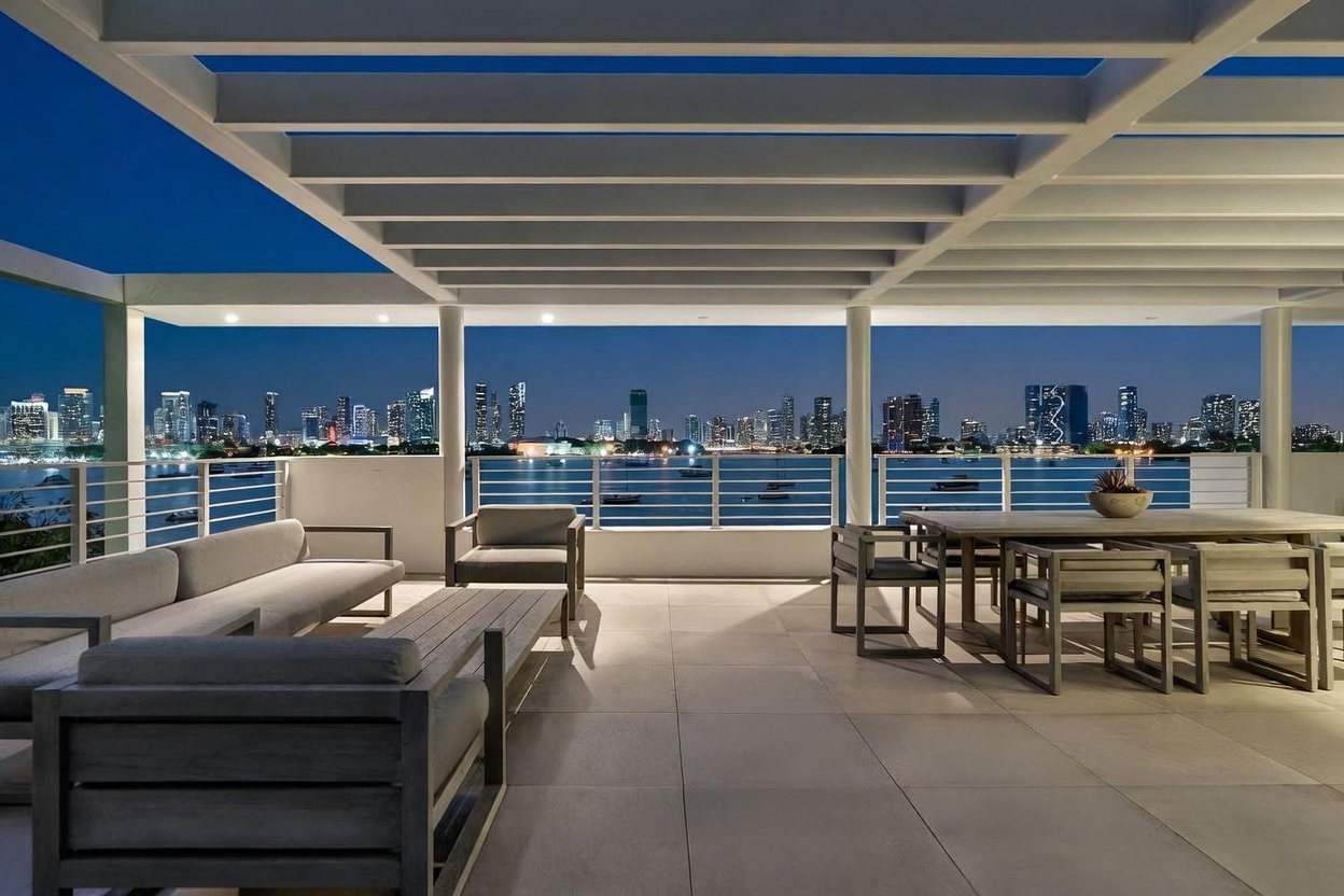 This expansive, covered balcony offers a sophisticated outdoor living space with a panoramic view of a city skyline across the water. The area is furnished with a modern sofa set and a large dining table, all set against a backdrop of a pergola-style ceiling and sleek metal railings. The perspective is a wide, eye-level shot that emphasizes the seamless transition between the luxurious terrace and the vibrant urban nightscape.
