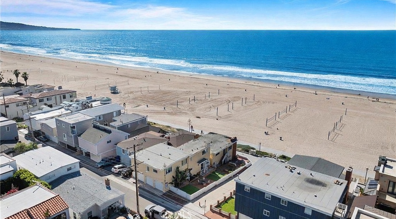This high-angle aerial shot captures a prime beachfront property situated directly along the sandy coastline. The perspective highlights the home's immediate proximity to the ocean, showcasing the surrounding neighborhood layout and the expansive beach area. A prominent location marker indicates the specific property, emphasizing its desirable coastal position and direct beach access.