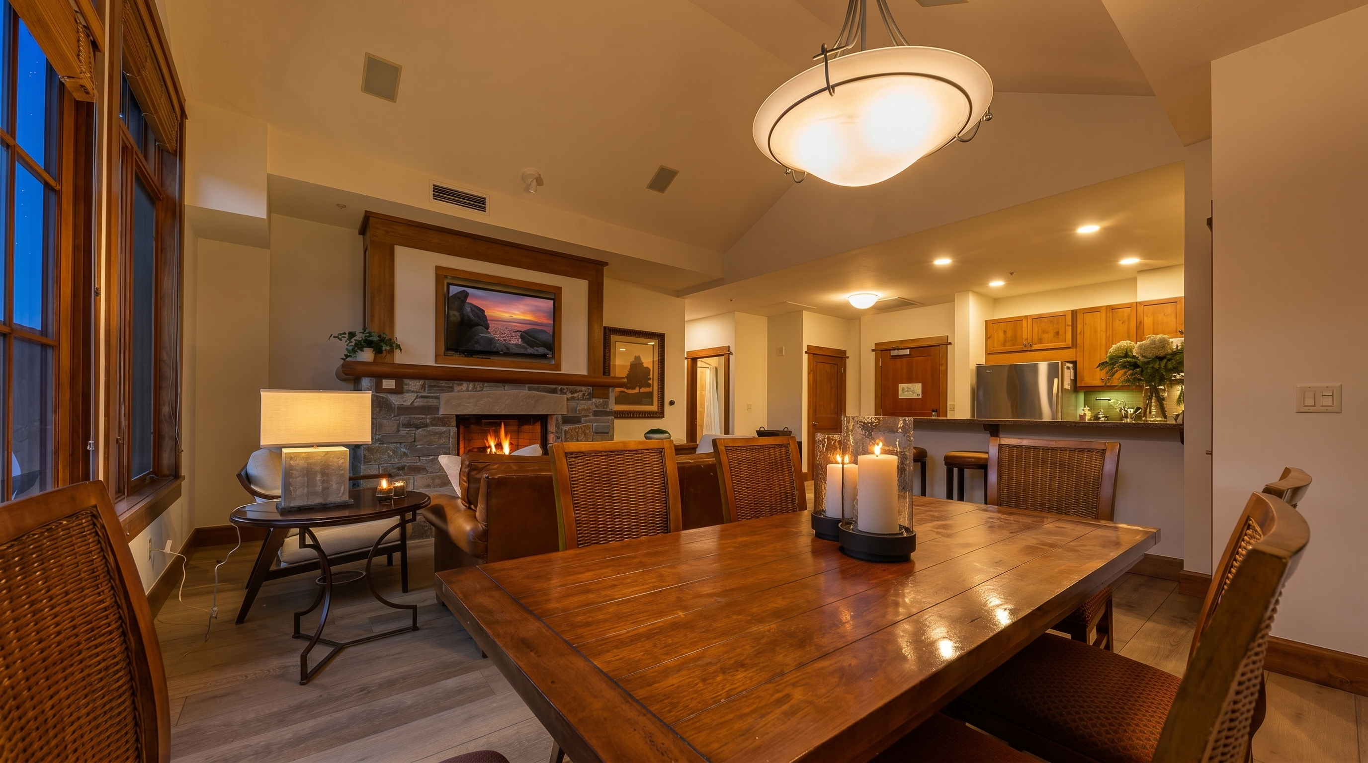 This open-concept living and dining area features a warm, rustic aesthetic with a large wooden dining table as the central focus. Beyond the dining space, a stone-clad chimney breast with a built-in television creates a cozy focal point for the adjacent living area, which includes a leather sofa and a side table with a lamp. The room is illuminated by a prominent modern pendant light fixture and recessed lighting, with light-toned wood flooring throughout and a view into the kitchen area in the background.