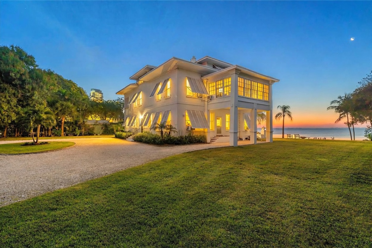 This elegant, multi-story white estate is captured at twilight, showcasing its prominent position along a serene waterfront. The home features classic architectural details, including numerous shuttered windows and a spacious, glass-enclosed sunroom that overlooks the calm water. A wide gravel driveway leads to the structure, set against a backdrop of lush greenery and a vibrant sunset sky.