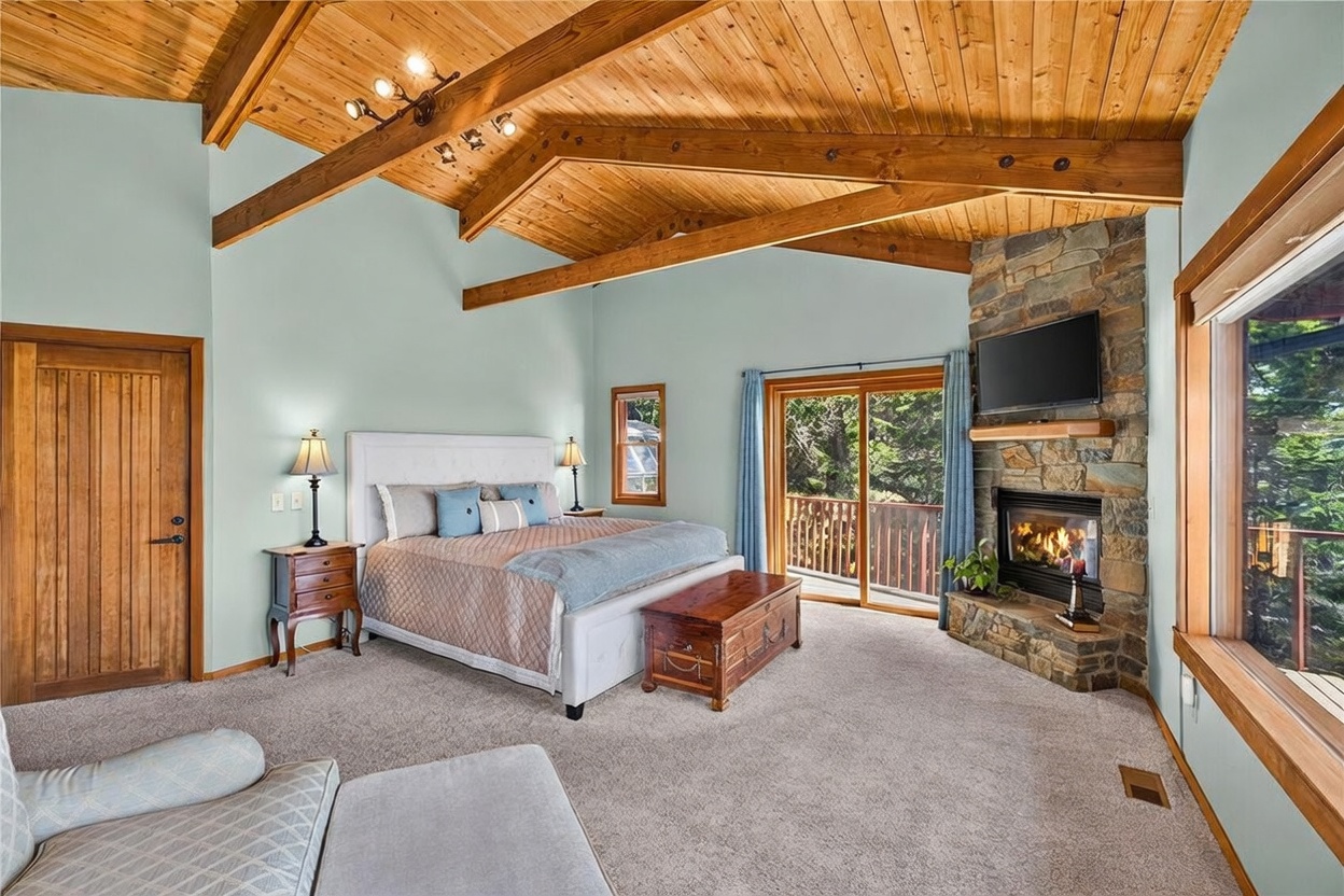 This spacious primary bedroom features a vaulted wood-paneled ceiling with exposed beams and a stone-clad chimney breast housing a fireplace. A large bed with a tufted headboard sits centrally, complemented by a wooden storage chest and matching nightstands. Large sliding glass doors provide access to an outdoor deck, filling the room with natural light and offering a view of the surrounding trees.