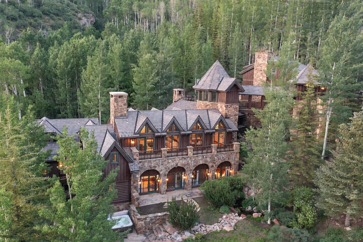This aerial perspective captures a grand, multi-story mountain estate nestled within a dense forest of aspen and pine trees. The home features a rustic architectural style with stone masonry, dark wood siding, and a complex roofline with multiple gables and a central tower. A stone patio on the lower level includes a hot tub, while a balcony above provides an elevated outdoor living space overlooking the wooded landscape.