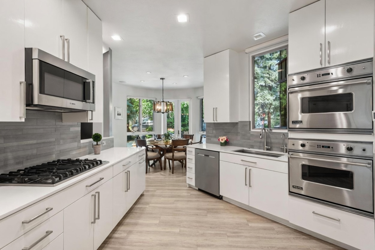 This bright, modern kitchen features crisp white cabinetry, stainless steel appliances, and light-toned wood-look flooring. The space is anchored by a gas range on the left and a double-oven unit on the right, with a seamless transition into an adjacent dining area illuminated by large windows. The clean lines and neutral color palette create an airy, inviting atmosphere perfect for contemporary living.