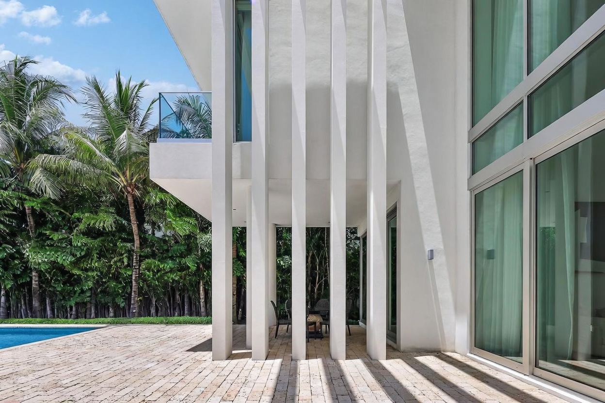 This modern outdoor living space features a covered patio area defined by striking, vertical white architectural columns that create a rhythmic play of light and shadow on the stone-paved deck. The area includes a small outdoor dining set and overlooks a pristine swimming pool, with lush tropical greenery providing a private backdrop. The clean lines and minimalist aesthetic emphasize a seamless transition between the indoor living space and the outdoor environment.
