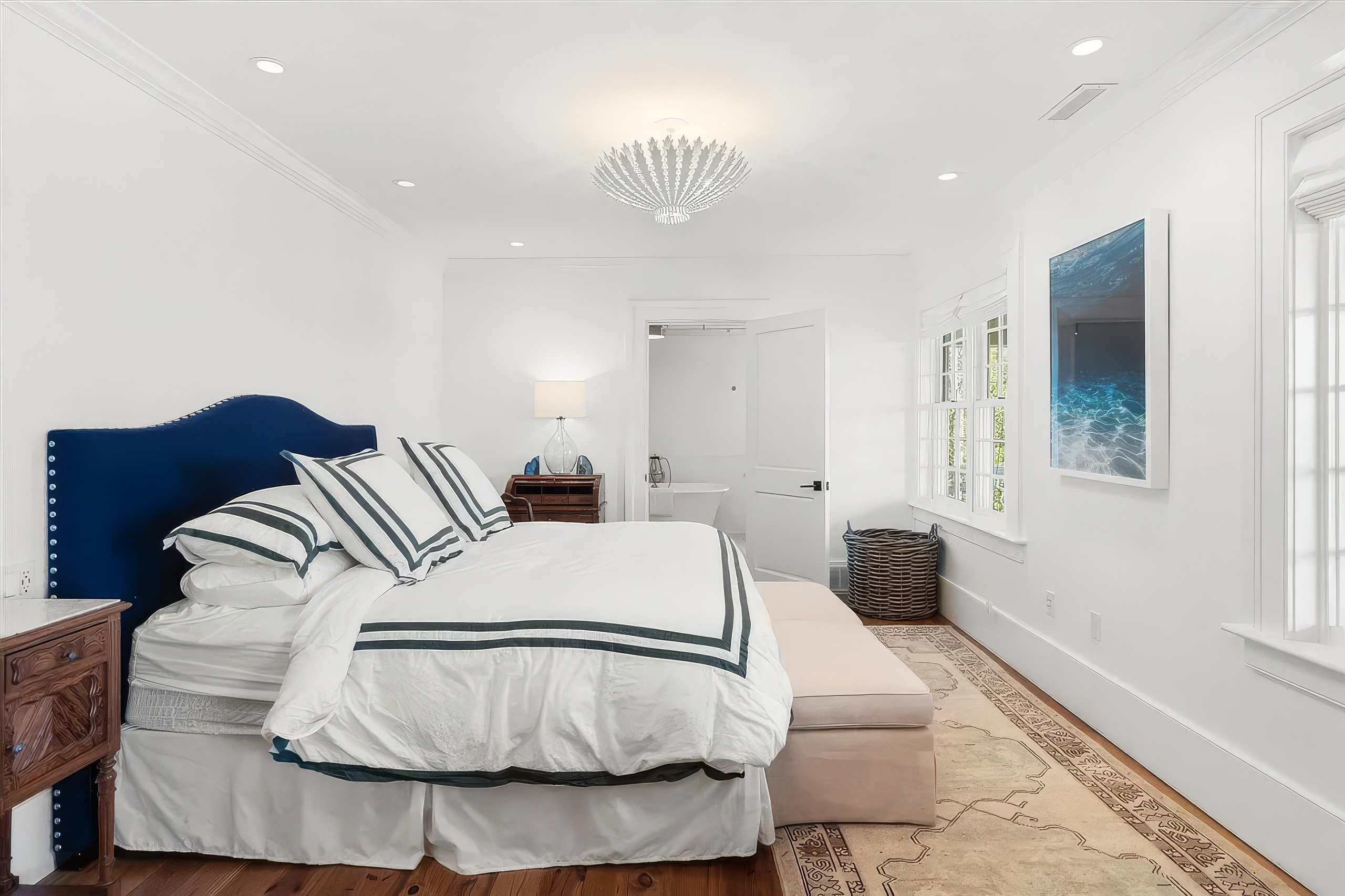 This primary bedroom features a bold navy blue upholstered headboard paired with crisp white bedding accented by dark striped pillows. A light-colored bench sits at the foot of the bed, resting on a patterned area rug that covers the wood flooring. The room is bright and airy, with white walls, a unique white chandelier, and a large piece of coastal-themed wall art, leading into an adjacent bathroom through an open doorway.