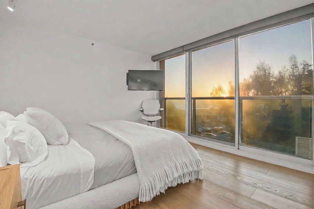 This bright and airy bedroom features a plush bed with white linens and a textured throw blanket, positioned next to expansive floor-to-ceiling windows that offer a scenic sunset view. A sleek, wall-mounted television and a modern white office chair are situated near the window, creating a functional and minimalist living space. The room is finished with light wood flooring, providing a warm and contemporary aesthetic.
