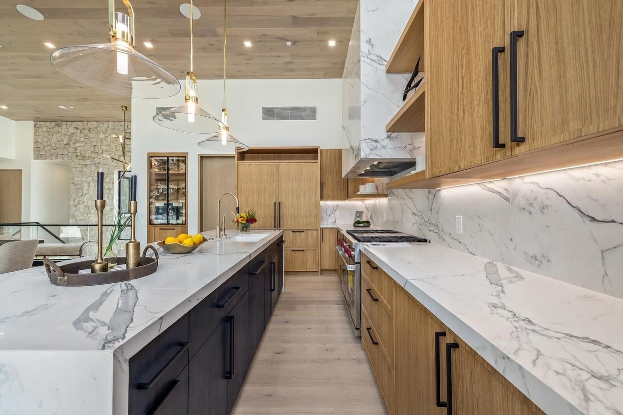 This modern kitchen features a striking contrast between dark cabinetry on the central island and light wood cabinetry along the perimeter. The space is anchored by a large island with a stone countertop, complemented by matching stone backsplashes and a full-height stone surround above the range. Three glass pendant lights hang from a light wood-paneled ceiling, creating a warm and sophisticated atmosphere.
