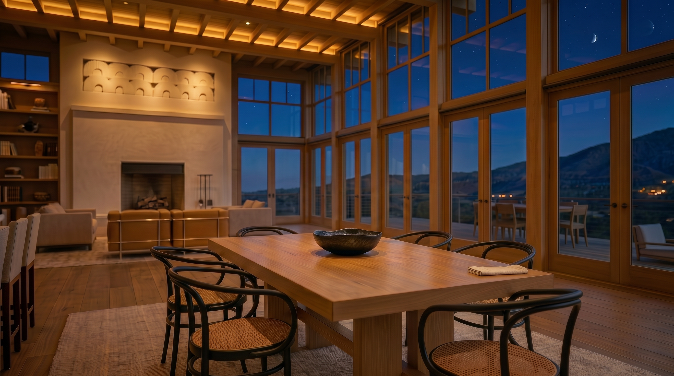 This spacious and sun-filled dining area features large, floor-to-ceiling windows that provide extensive natural light and scenic views. The room is anchored by a sturdy, minimalist wooden dining table, and its high beamed ceiling adds a touch of rustic sophistication. With an open floor plan leading toward a cozy fireplace area, this space is designed for comfortable gathering and entertaining in a modern, elegant setting.