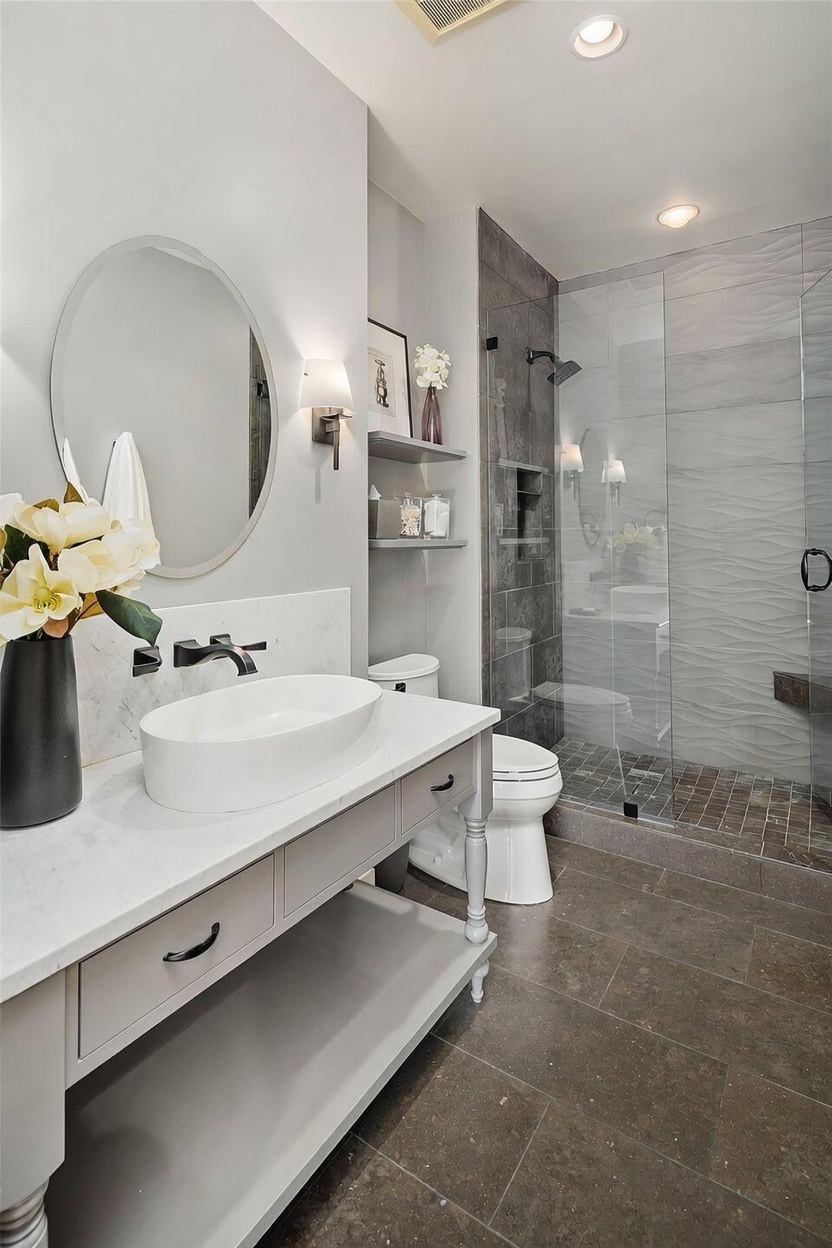 This modern bathroom features a sleek vanity with a vessel sink, a round mirror, and wall-mounted lighting. A glass-enclosed shower with textured wall tiles and a dark stone floor sits adjacent to a white toilet and open shelving. The neutral color palette and clean lines create a sophisticated and contemporary atmosphere.