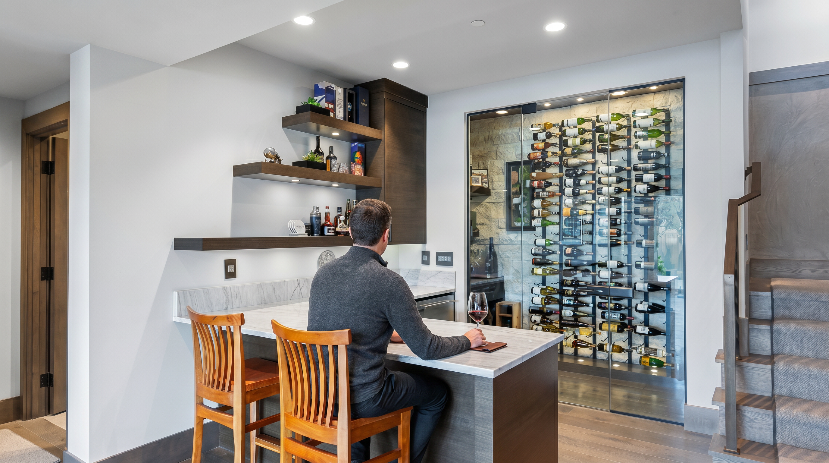 This sophisticated home bar area features a sleek, dark wood counter with a stone top and two wooden stools, complemented by open shelving for spirits and glassware. Adjacent to the bar is a climate-controlled wine cellar enclosed by floor-to-ceiling glass, showcasing an extensive collection of bottles on metal racks against a textured stone wall. The space is illuminated by recessed lighting and provides a refined, modern atmosphere perfect for entertaining.