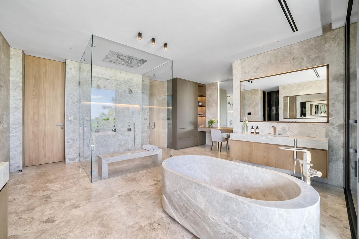 This luxurious primary bathroom features a large, freestanding stone soaking tub as the centerpiece, complemented by a spacious walk-in glass shower with a built-in stone bench. A floating vanity with a large mirror and a separate makeup vanity area with a chair create a functional and elegant layout. The room is finished with neutral stone surfaces throughout, providing a sophisticated and serene atmosphere.