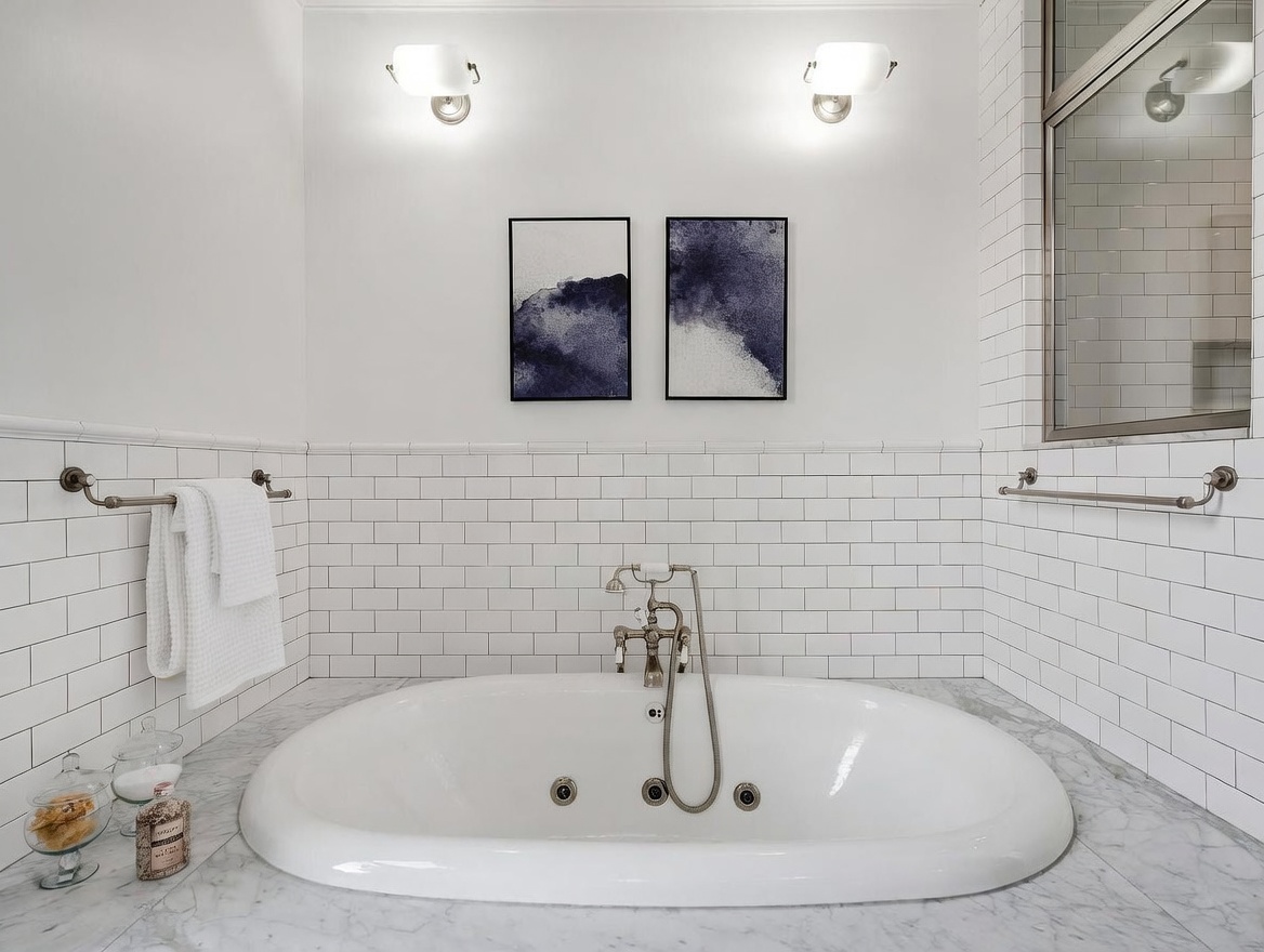 This elegant bathroom features a deep soaking tub set into a stone-topped deck, centered against a wall of classic white subway tile. Two abstract blue-toned artworks hang symmetrically above the tub, flanked by wall-mounted sconces that provide soft, warm lighting. The space is finished with polished metal fixtures and a towel rack, creating a clean, spa-like atmosphere.