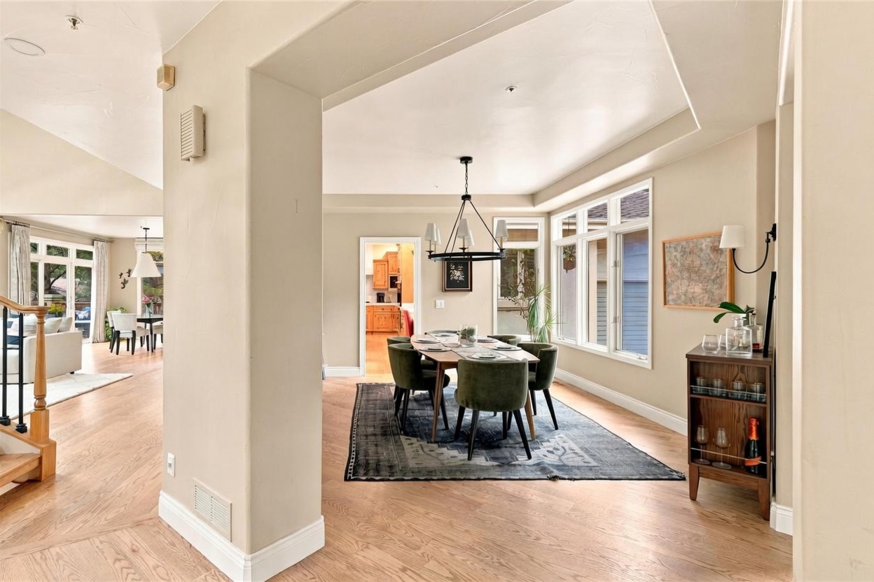 This bright and open dining area features a central wooden table surrounded by six upholstered green chairs, set atop a large, dark patterned area rug. The space is illuminated by a modern black chandelier and benefits from natural light streaming through a series of large windows along the side wall. A wooden bar cabinet stands to the right, while a glimpse into the adjacent living room and kitchen adds to the home's cohesive, flowing floor plan.