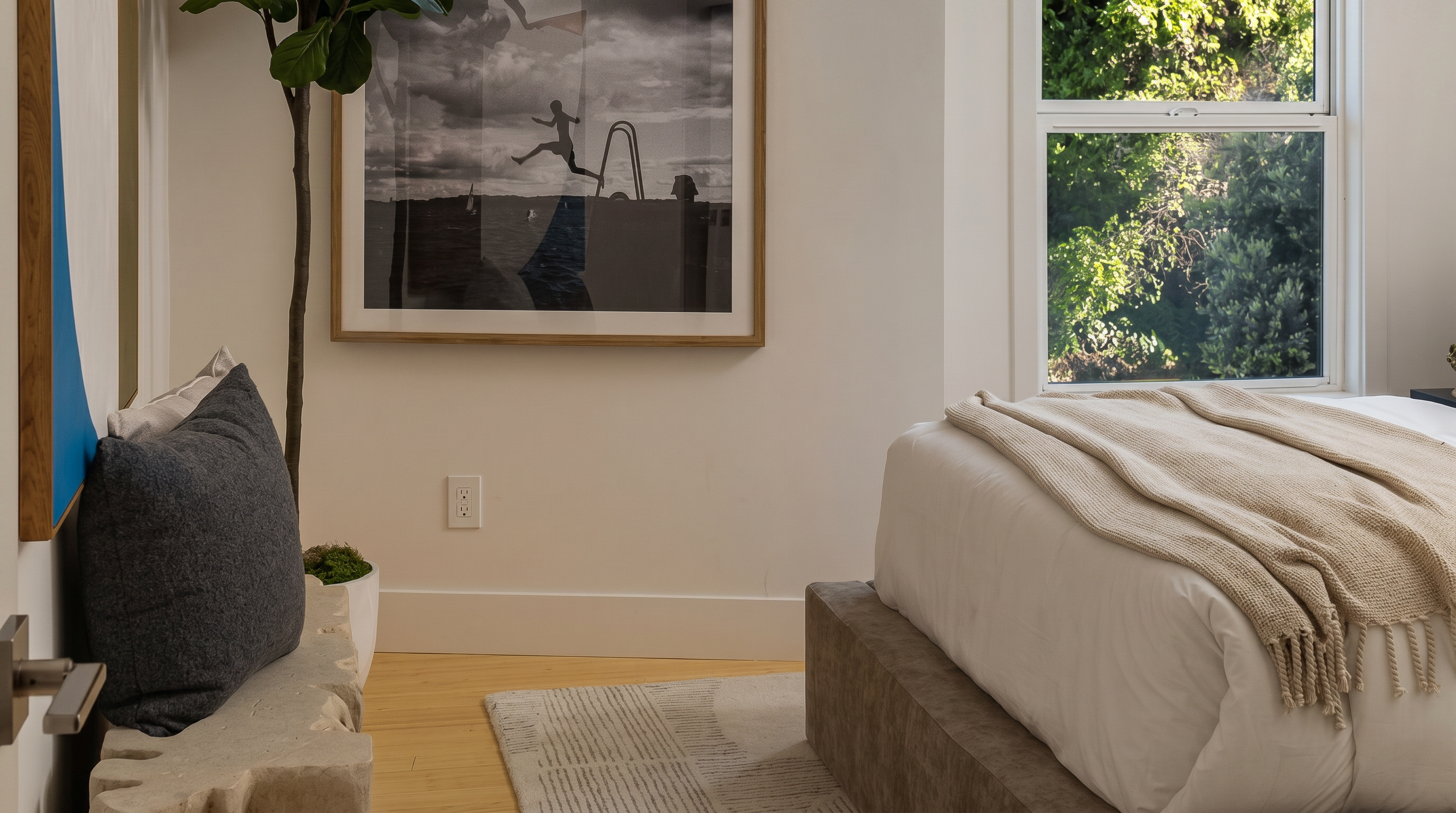 This tranquil primary bedroom features a modern platform bed with a neutral textured throw, creating an inviting and minimalist aesthetic. The space is accented by a sculptural stone decor element, a large framed black-and-white art piece depicting a diver, and warm wood flooring. Natural light streams in through a side window, highlighting the clean lines and sophisticated, relaxed atmosphere of the room.