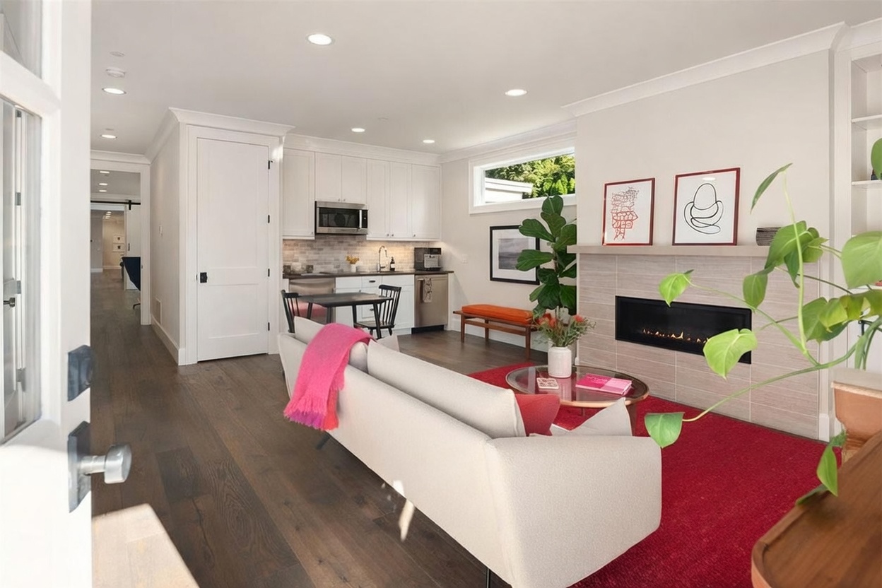 This open-concept living space features a modern aesthetic with dark wood flooring and a bright, neutral color palette. The room is anchored by a fireplace with a sleek, horizontal insert and a light-toned surround, complemented by a vibrant red area rug and a contemporary sofa. In the background, a compact kitchen area with white cabinetry and a small dining table creates a seamless flow, while a large potted plant adds a touch of greenery to the bright, airy atmosphere.