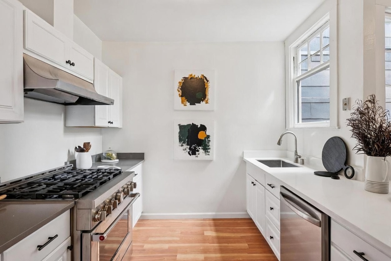 This bright, galley-style kitchen features crisp white cabinetry paired with dark stone countertops and stainless steel appliances, including a professional-grade gas range. Two abstract art pieces hang on the central wall, adding a modern touch to the clean, minimalist aesthetic. The space is illuminated by natural light from a side window and features warm-toned wood flooring.