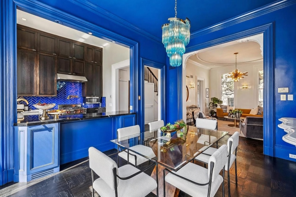 This vibrant dining area features bold blue walls and trim, centered around a glass-topped table with six modern upholstered chairs. A striking blue tiered chandelier hangs above the table, while the space opens directly into a sleek kitchen with dark cabinetry and a blue herringbone backsplash. The perspective looks through the dining space into an adjacent living room, showcasing an open-concept layout with rich dark wood flooring throughout.