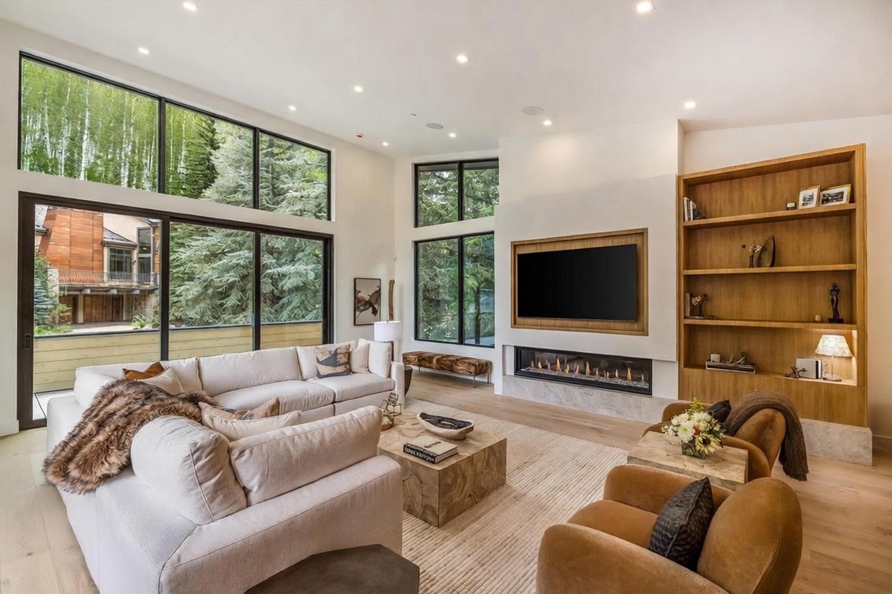 This spacious living room features a contemporary design with a large, light-colored sectional sofa and two plush, warm-toned armchairs arranged around a textured wooden coffee table. A prominent fireplace with a stone base and a recessed television above serves as the focal point, complemented by a tall, built-in wooden shelving unit. Large windows provide an expansive view of the surrounding forest, filling the room with natural light and creating a seamless connection to the outdoors.