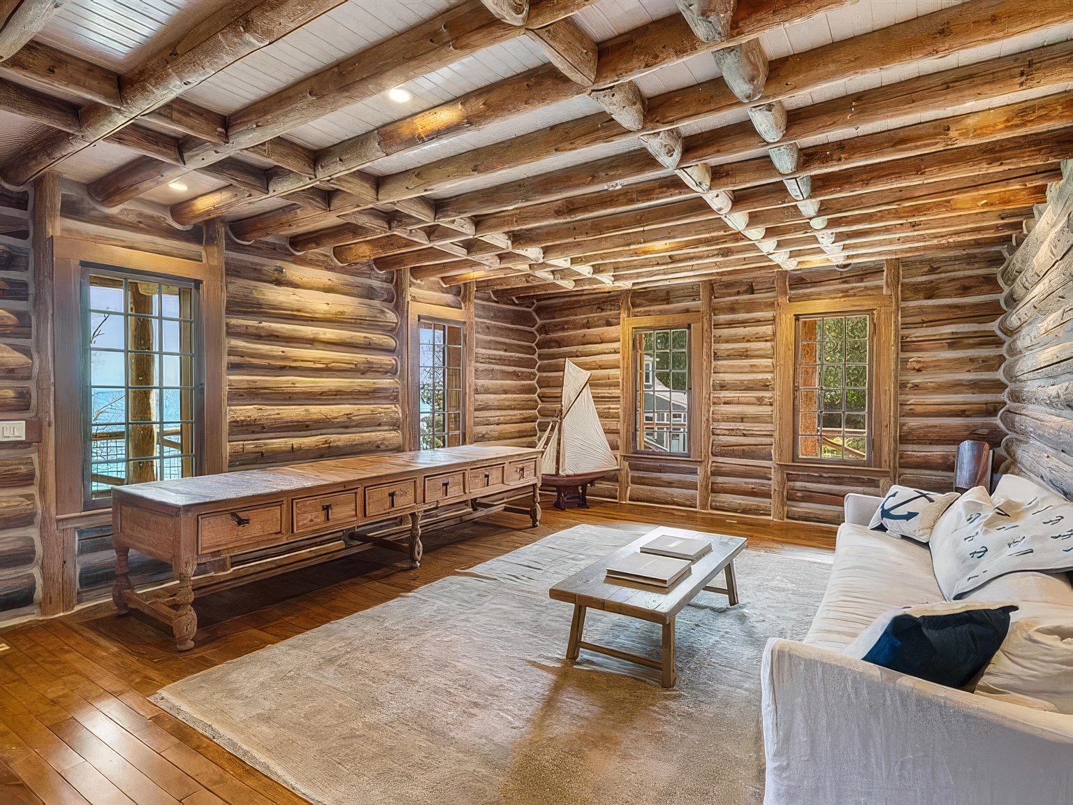 This rustic living room features exposed log walls and a beamed ceiling, creating a cozy, cabin-like atmosphere. The space is furnished with a long wooden console table, a low coffee table, and a comfortable sofa adorned with nautical-themed pillows. Large windows provide ample natural light and views of the outdoors, while a model sailboat serves as a decorative focal point in the corner.
