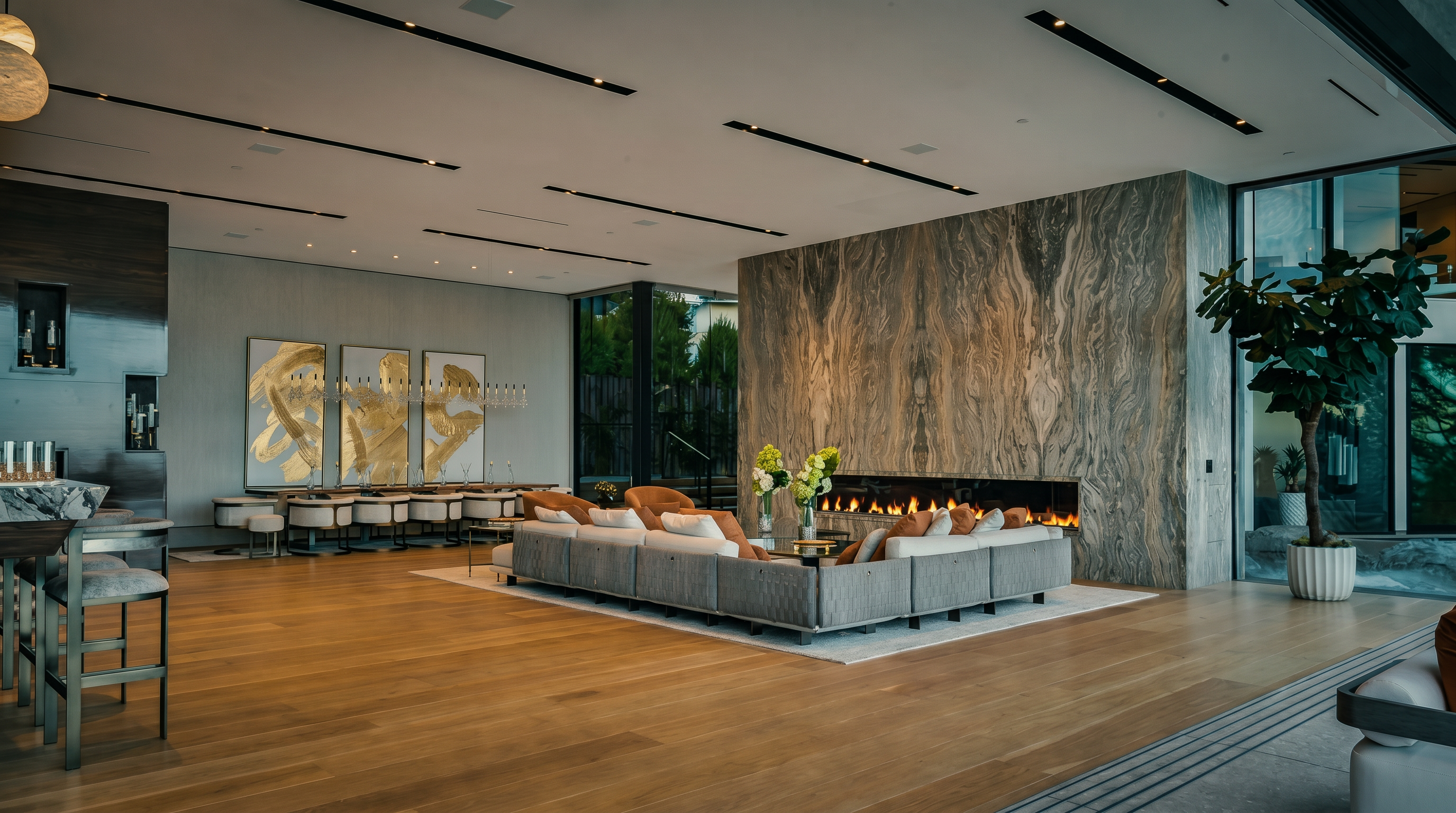 This expansive, open-concept living room features a striking floor-to-ceiling stone fireplace wall and sleek, recessed linear lighting across the high ceiling. A large, modular grey sectional sofa anchors the space, complemented by light hardwood flooring and a sophisticated dining area in the background. The room exudes a luxurious, modern aesthetic with floor-to-ceiling windows that invite natural light and provide a seamless transition to the outdoors.