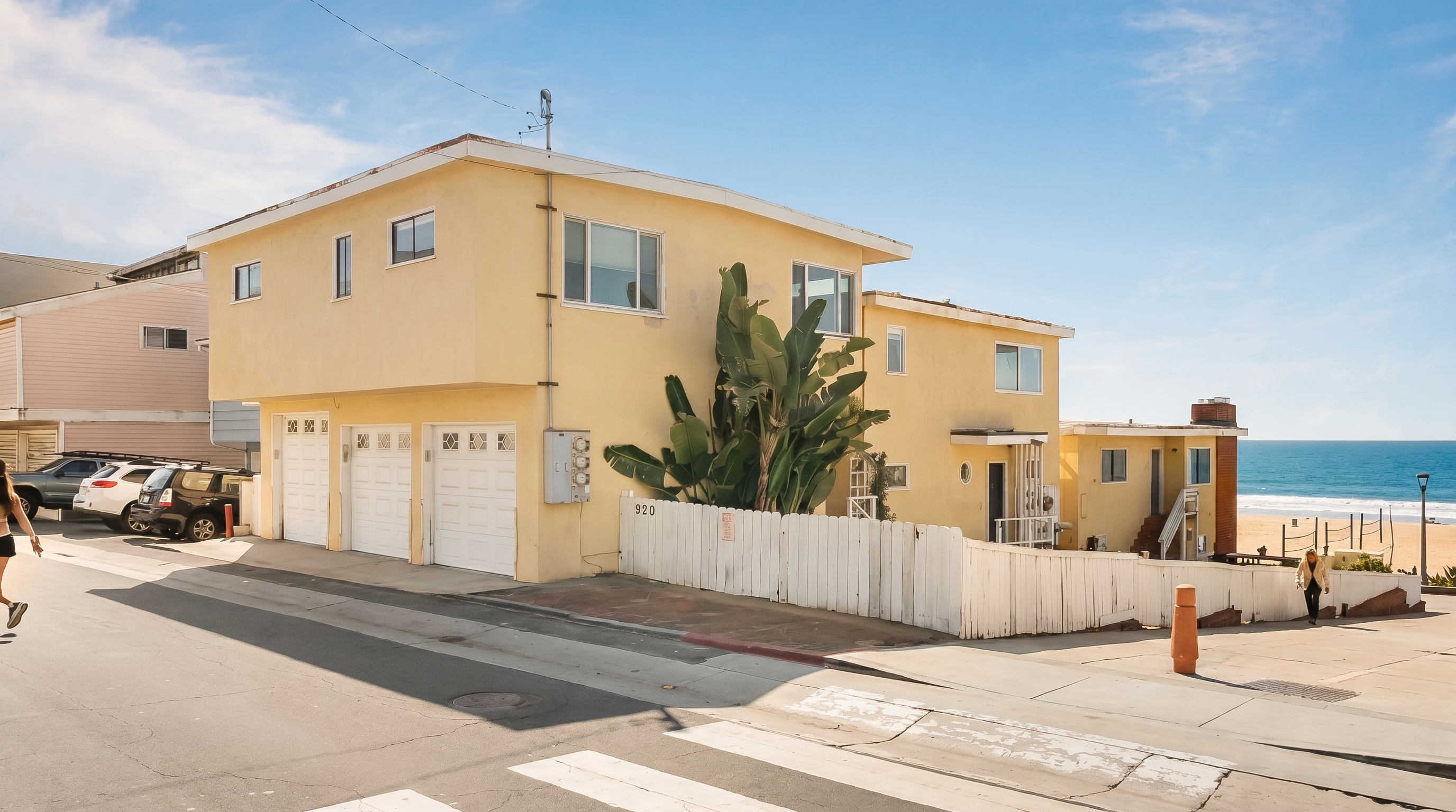 This exterior view captures a multi-level, light yellow residential building situated directly adjacent to a sandy beach and the ocean. The property features a three-car garage on the street level, a white picket fence enclosing a side area, and a prominent, mature tropical plant near the entrance. The perspective is taken from the street, highlighting the home's prime coastal location and its proximity to the shoreline.